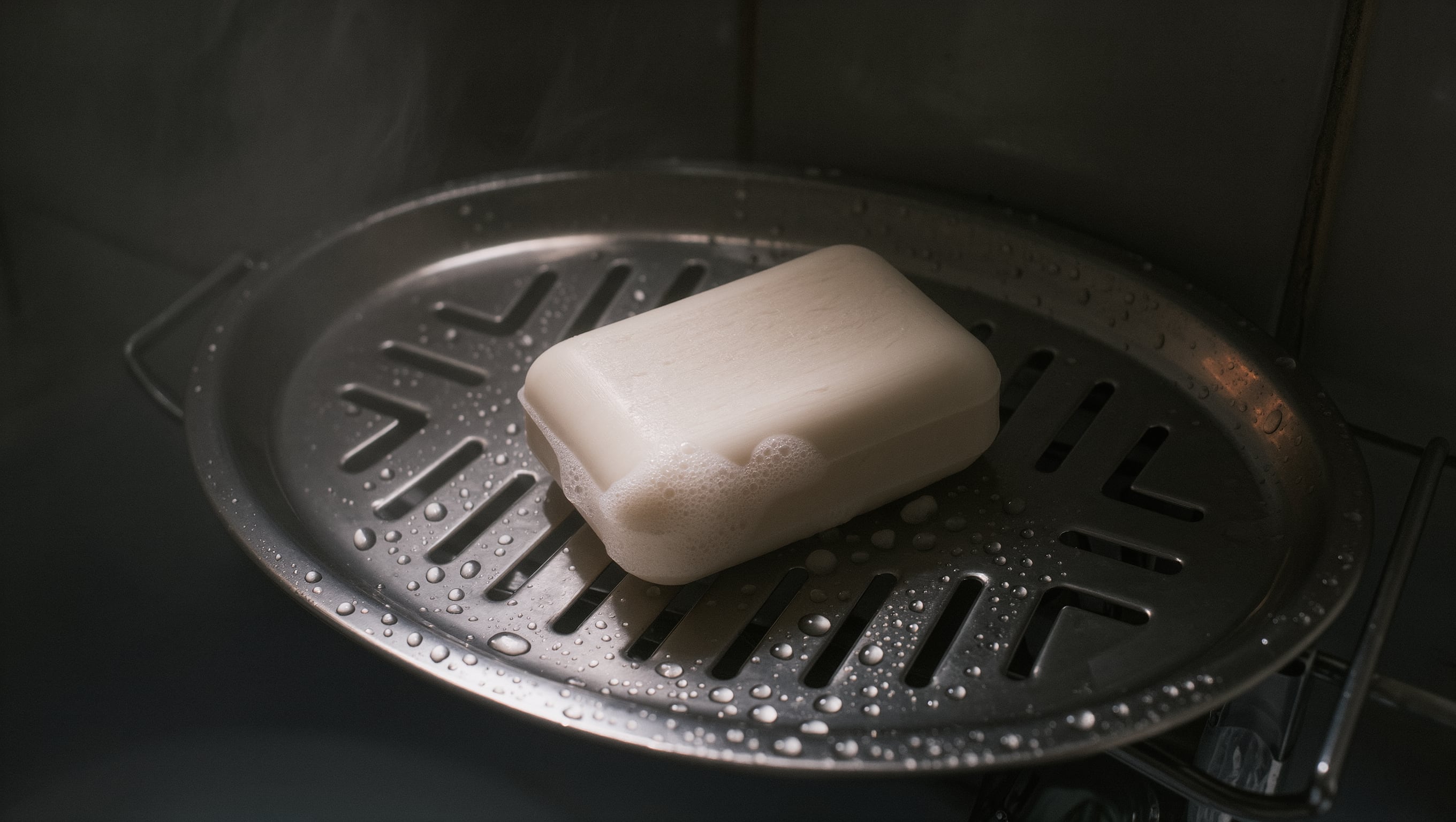 Ein angebrauchtes festes Shampoostück liegt auf einer Ablaufschale aus Metall, Wassertropfen und Schaumreste im dunklen Bad